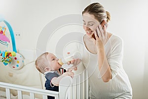 Portrait of smiling young businesswoman talking by phone and feeding her little baby son