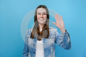 Portrait of smiling young caucasian brunette woman waving-hand his, positive looking at camera, dressed in denim jacket