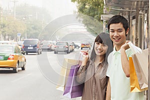 Portrait of smiling young couple carrying colorful shopping bags and waiting for the bus at the bus stop, Beijing, China