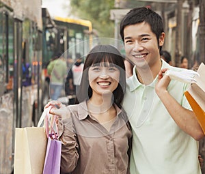 Portrait of smiling young couple carrying colorful shopping bags and waiting for the bus at the bus stop, Beijing, China