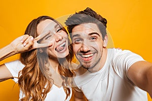 Portrait of smiling young couple man and woman taking selfie photo and showing peace sign, isolated over yellow background