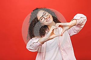 Portrait of a smiling young curly woman showing heart gesture with two hands and looking at camera isolated over red background