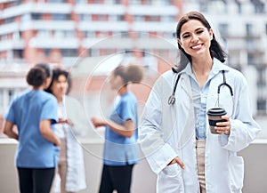 Portrait of smiling young doctor enjoying a cup of coffee outside with her colleagues in the background. Happy medical