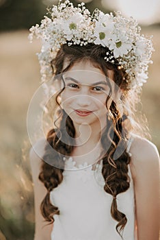 Portrait of a smiling young girl in a white dress and a floral wreath on her hair with braids in the summer field