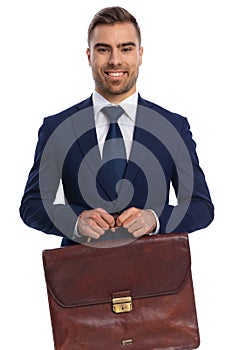 portrait of smiling young guy in navy blue suit proudly holding suitcase
