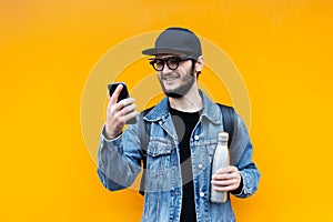 Portrait of smiling young hipster guy, holding steel thermo eco bottle for water and looking in smartphone, on orange background.