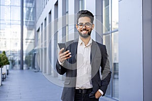 Portrait of a smiling young Indian businessman in a suit standing outside an office building, holding his hand in his