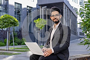 Portrait of a smiling young Indian man sitting in a business suit, wearing headphones, holding a laptop on his lap