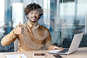 Portrait of a smiling young Indian man sitting at an office desk with a laptop, looking at the camera and showing a