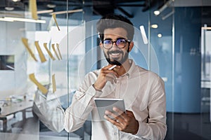 Portrait of a smiling young Indian man standing in the office near a glass board with notes, holding a planchet in his