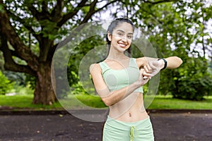 Portrait of a smiling young Indian woman in sportswear jogging, standing in a park, checking the time on a smart watch