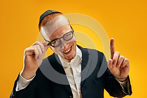 Portrait of smiling young Jewish man in yarmulke, glasses and suit raising finger up against yellow studio background