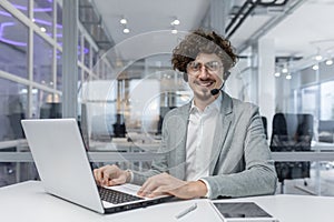 Portrait of a smiling young man in a business suit sitting at a desk in a headset working in the office on a laptop. He