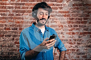 Portrait of smiling young man with earphones and cell phone. Casual wear and brick wall of background