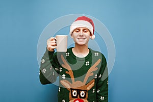 Portrait of smiling young man in green warm sweater and hat of santa claus, standing against blue background with cup hot drink in