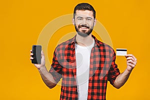 Portrait of a smiling young man holding blank screen mobile phone and showing credit card isolated over yellow background