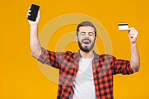 Portrait of a smiling young man holding blank screen mobile phone and showing credit card isolated over yellow background