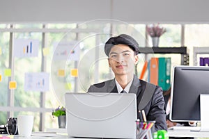 Portrait of smiling young man sitting at his desk in the office, Businessman in suit working with laptop in office