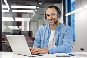 Portrait of a smiling young man sitting in the office with a laptop in headphones and looking confidently into the