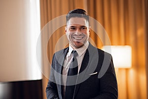 A portrait of a smiling young man in a suit, white shirt and a black tie standing in the middle of a hotel room