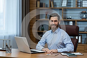 Portrait of a smiling young man wearing headphones sitting and working on a laptop in the office and looking confidently