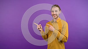 Portrait of smiling young man in yellow bright shirt pointing to space on isolated purple background. 4K