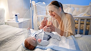 Portrait of smiling young mother smiling at her little baby son lying on bed and holding his little feet. Happy