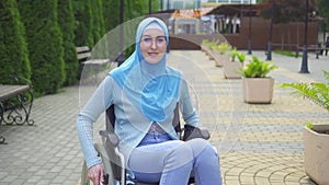 Portrait smiling young muslim woman disabled in traditional scarf sitting in a wheelchair in the park