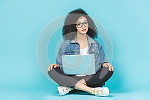 Portrait of smiling young afro american woman using laptop while sitting on a floor with legs crossed isolated over blue