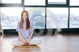 Portrait of smiling young relaxed woman meditating sitting on floor in lotus position on background of window in bright