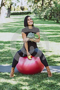 Portrait of smiling young tanned woman in the park holding an apple and a bouquet of greens with broccoli, diet, healthy eating.