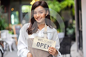 Portrait of a smiling young woman in apron holding open sign board while standing at the cafe. The coffee shop is raising the sign