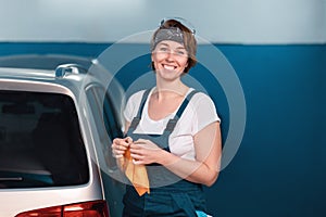 Portrait of smiling young woman in blue coveralls. Car and workshop at the background. Work at auto repairshop and