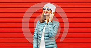 Portrait of smiling young woman calling on smartphone wearing jacket and hat looking away on red background