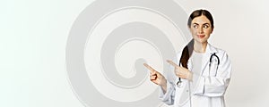 Portrait of smiling young woman doctor, healthcare medical worker, pointing fingers left, showing clinic promo, logo or