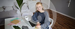 Portrait of smiling young woman, female student, doing distance learning course, using laptop, studying at home, writing