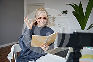 Portrait of smiling young woman, holding notebook, showing okay, ok sign, looking at digital camera, recording video