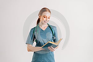Portrait of smiling young woman intern in green uniform with stethoscope reading medical book standing on white isolated