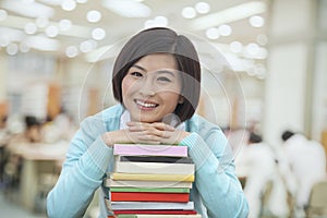 Portrait of Smiling Young Woman in Library Leaning on a Stack of Books, Looking At Camera