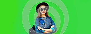 Portrait of smiling young woman model wearing black round hat, denim jacket and backpack on green background