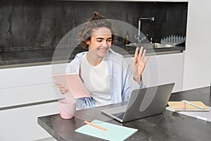 Portrait of smiling young woman says hi, waves at laptop camera, video chats with her tutor. Teacher connects to a