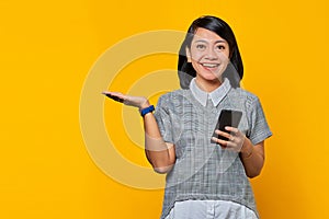 Portrait of smiling young woman showing copy space in palm and holding mobile phone on yellow background