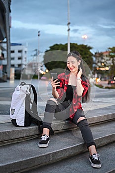 Smiling young woman sitting on stairs in the city at night and using smart phone.