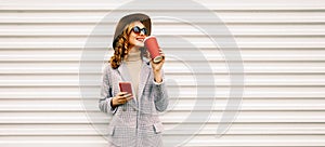 Portrait of smiling young woman with smartphone drinking coffee looking away wearing coat jacket, round hat on white background