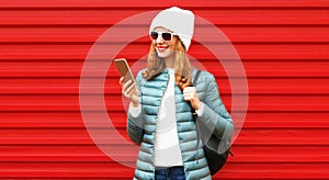 Portrait of smiling young woman with smartphone wearing jacket, backpack, white hat on red background