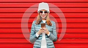Portrait of smiling young woman with smartphone wearing jacket, white hat on red background