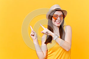 Portrait of smiling young woman in straw summer hat, orange glasses pointing index fingers aside copy space isolated on