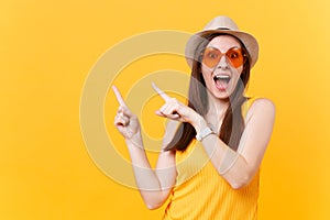 Portrait of smiling young woman in straw summer hat, orange glasses pointing index fingers aside copy space isolated on