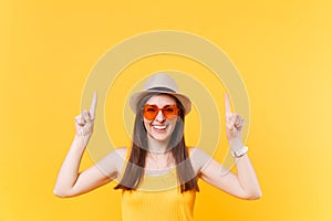 Portrait of smiling young woman in straw summer hat, orange glasses pointing index fingers up on copy space isolated on