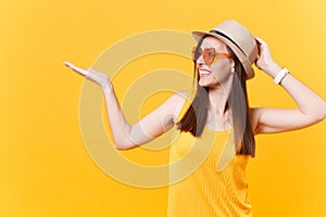 Portrait of smiling young woman in straw summer hat, orange glasses pointing right hand aside copy space isolated on